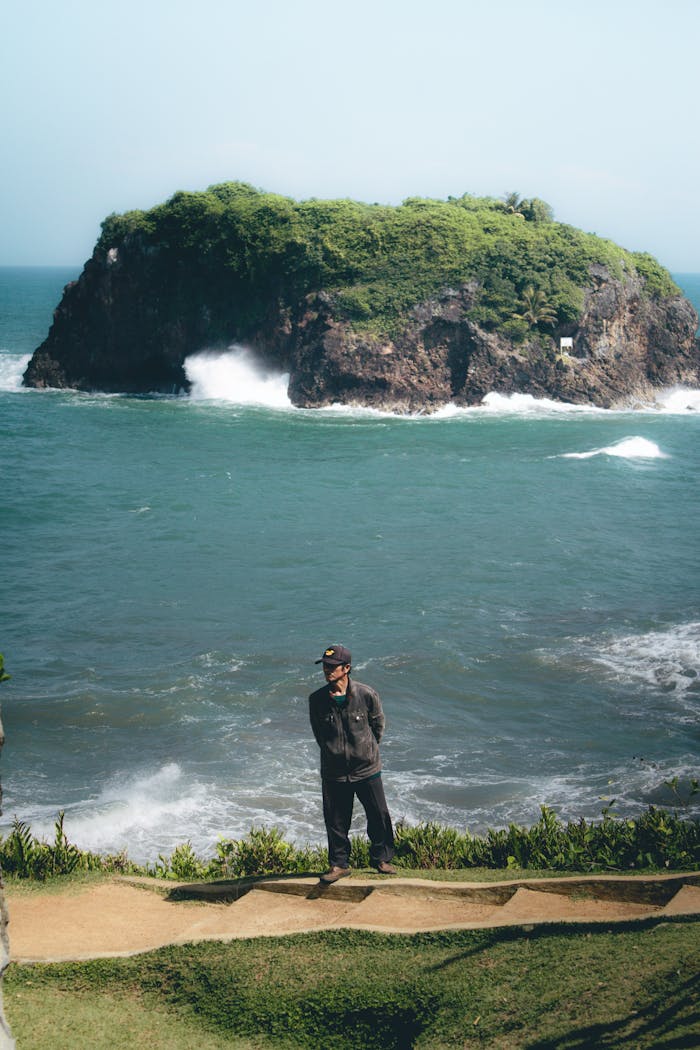 A man stands on a cliff overlooking the ocean with a lush island in the background, located in West Java, Indonesia.